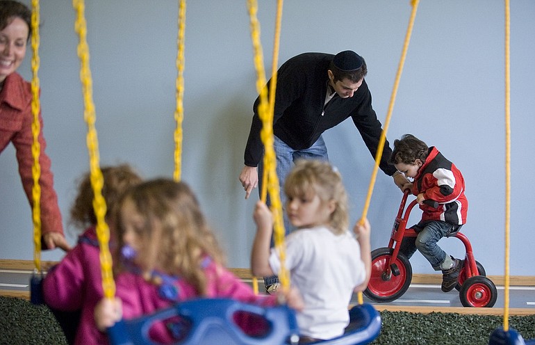 Three-year-old Samuel Goyzman, background, gets help riding a tricycle from his father, Joseph Goyzman, while other youngsters play on an indoor swing set during the dedication ceremony for the Gan Garrett Jewish Preschool at the Chabad Jewish Center in the Sifton neighborhood Sunday.