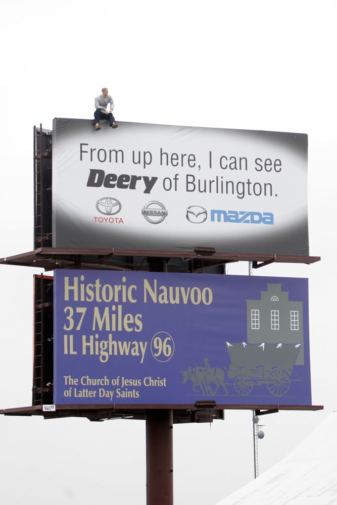 A mannequin sits Thursday on top of a Deery Brothers billboard on the southbound side of Highway 61 near Mediapolis, Iowa. The mannequin is so lifelike that it has fooled many drivers into calling 911, believing their is a real person on top of the sign.