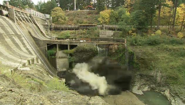 The first dust and smoke emerges from the base of Condit Dam as a 700-pound charge of TNT detonates, clearing the final section of tunnel underneath the dam.