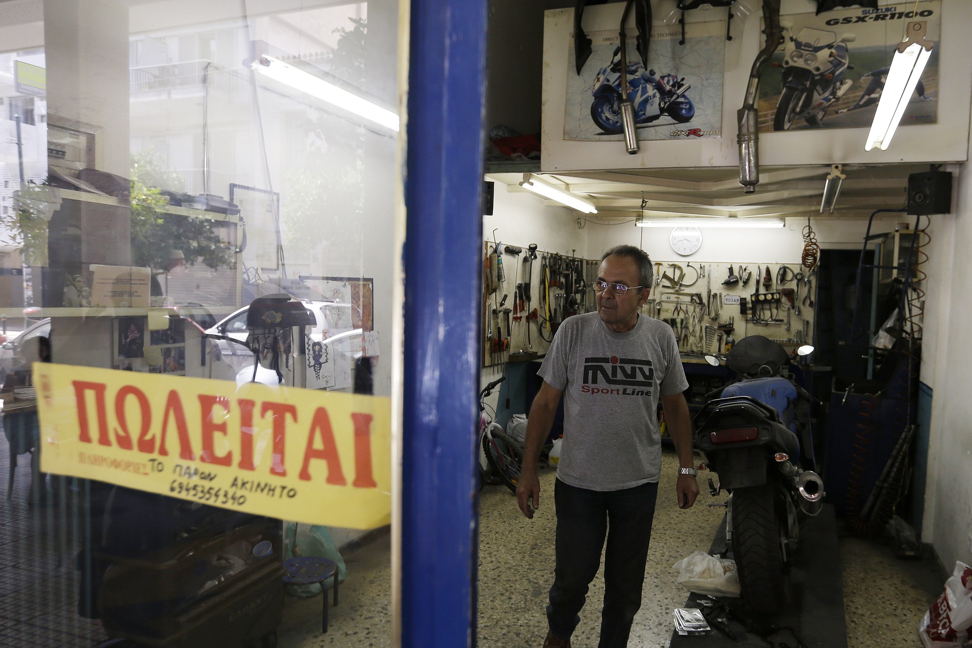 Mechanic Giorgos Prasinoudis steps out of his closed motorcycle repair shop, as a 'For Sale' sign is posted on the front window, in Athens, on Wednesday, July 22, 2015. Uncertainty over Greece's bailout and recent capital controls have led to a new spate of store closures in Greek capital.