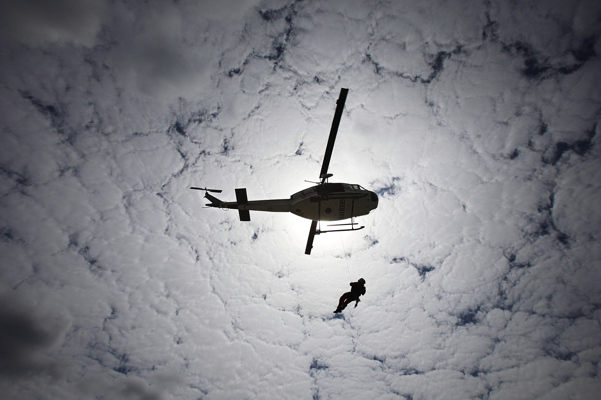 A Search and Rescue volunteer glides down to the ground on a line from the Sheriff Department's Huey helicopter with cloud cover above as part of an August 2012 training exercise at Taylor's Landing Search and Rescue Facility in Snohomish.