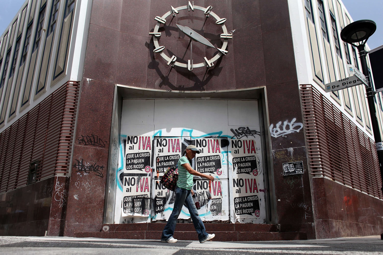 A woman walks in front of a closed down bank in the neighborhood of Rio Piedras in San Juan, Puerto Rico, Monday, June 29, 2015. The bills on the closed bank doors read in Spanish &quot;No to the value added tax. Let the rich pay for the crisis.&quot; International economists released a critical report on Puerto Rico's economy Monday on the heels of the governor's warning that the island can't pay its $72 billion public debt.