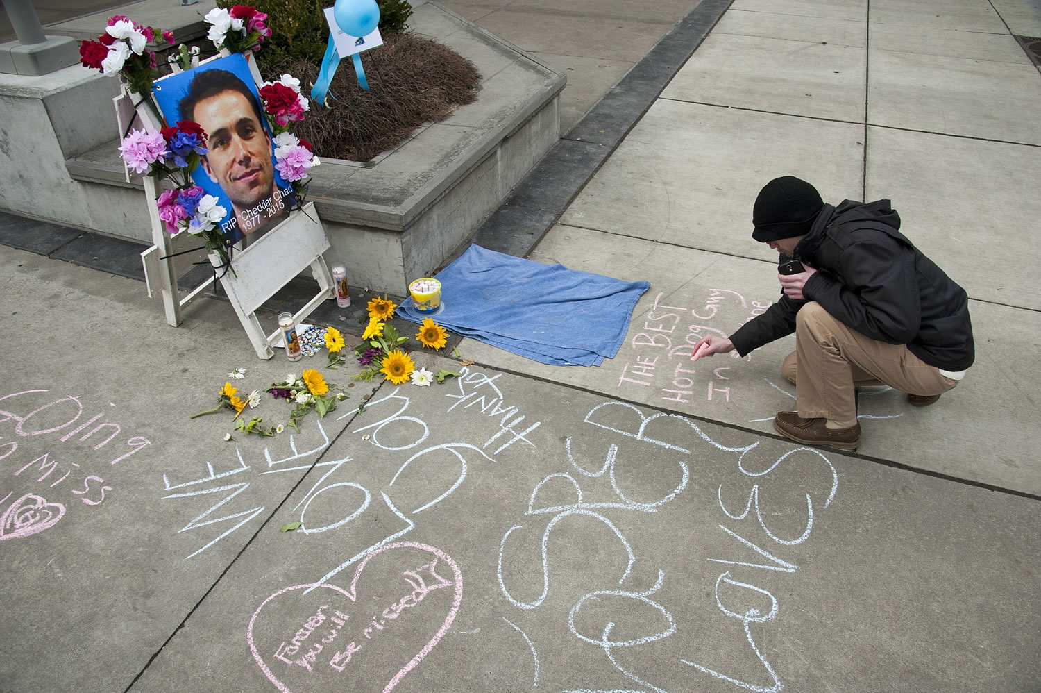Casey Devine writes a chalk message Wednesday at a sidewalk memorial to hot dog vendor Chad Rattray in Spokane. Rattray had been a fixture on the corner for the past 13 years.