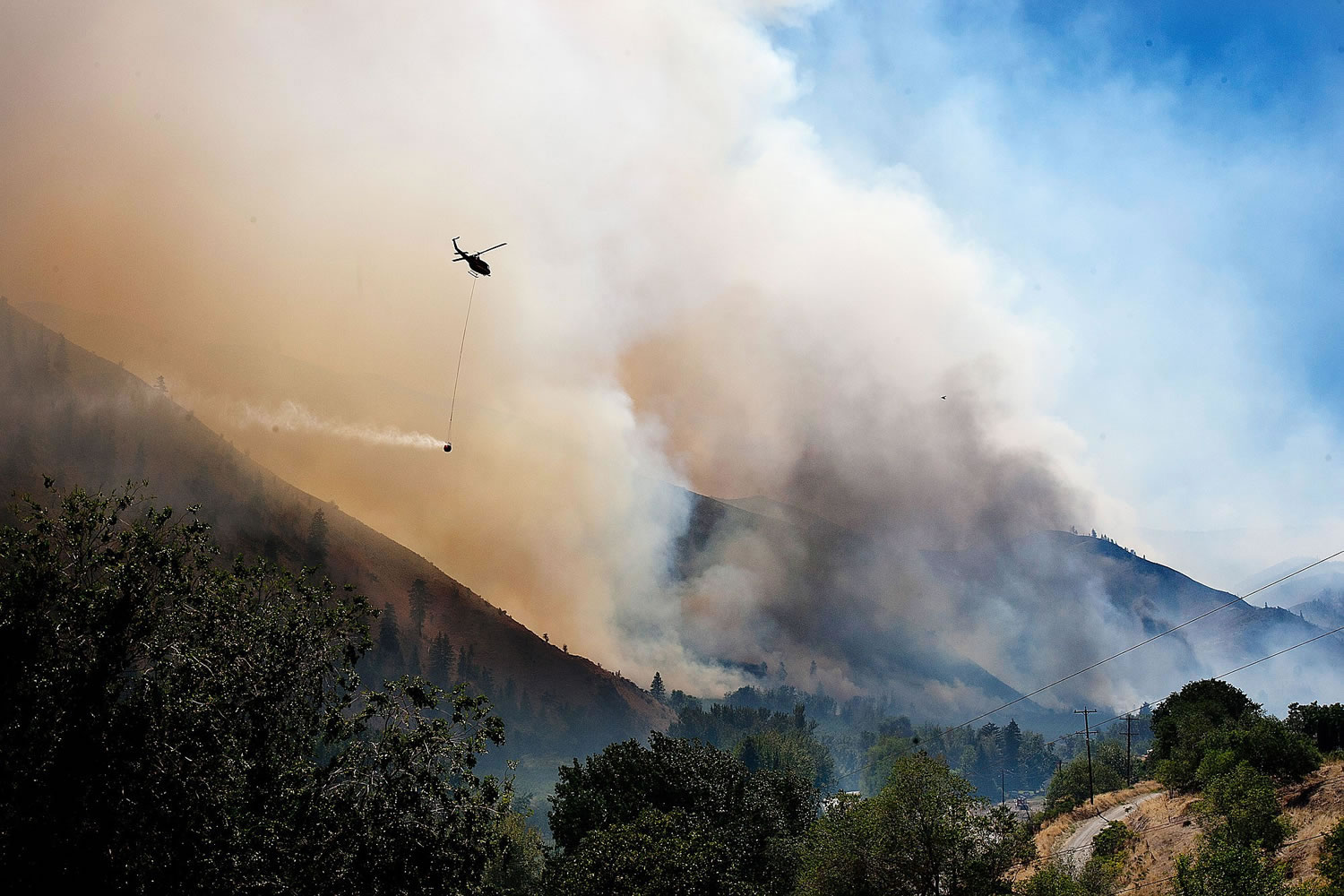 As smoke and fire blow through the lower Entiat Valley near Entiat on Wednesday afternoon, a helicopter with a bucket of water flies to the fire to drop its load.