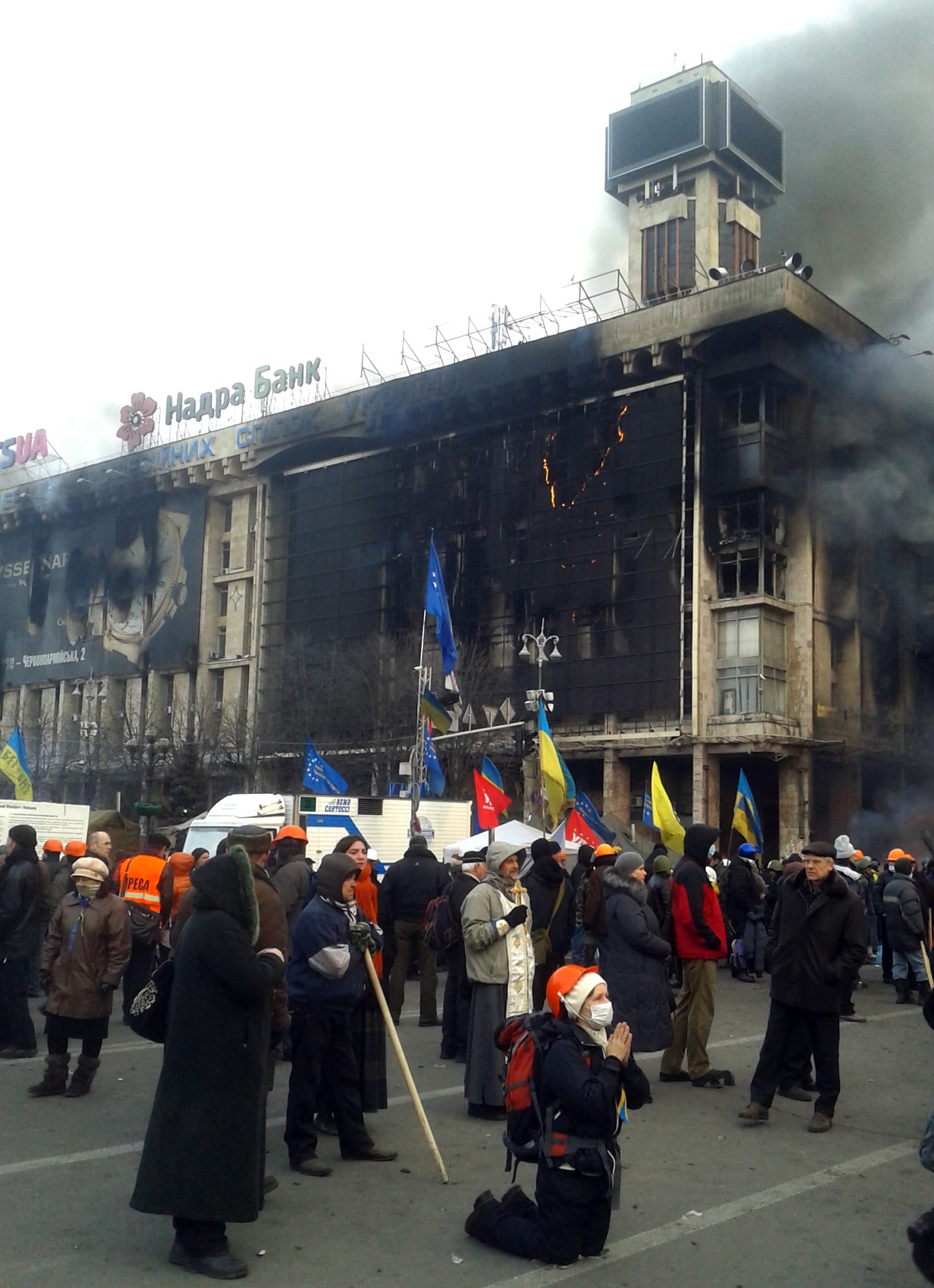 A young woman wearing a safety helmet kneels in prayer as smoke rises from a burning building on a corner of Independence Square in Kiev, Ukraine.