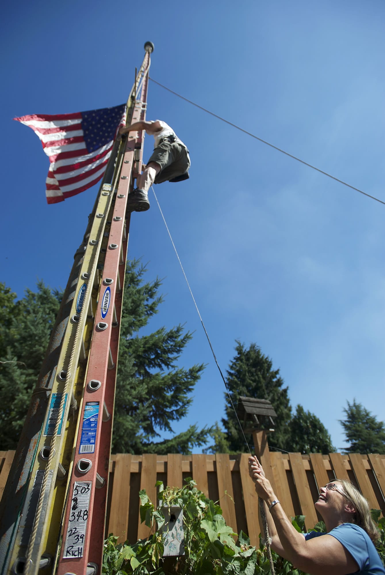 Pam McNamara hoists the flag in the patio of her home near Crown Park in Camas as Rich Ullsmith ascends a ladder to ensure the flagpole system he built is working smoothly.