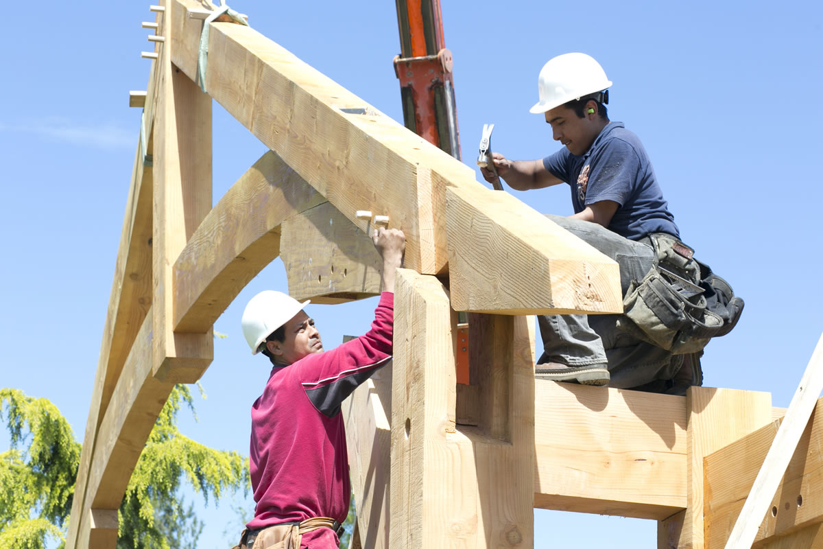 Suspended by heavy cable attached to a reach forklift's beefy hooks, a huge truss is gently lowered into place as a worker from Arrow Timber Framing hammers the tenon into a corresponding mortise in the support beam.