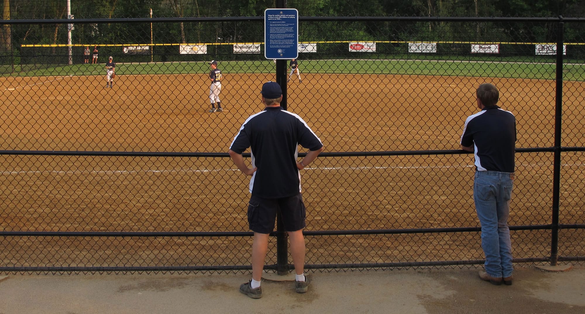 Coaches and parents watch a youth baseball game near a sign on a fence about parent behavior in Buffalo Grove, Ill. on June 10.
