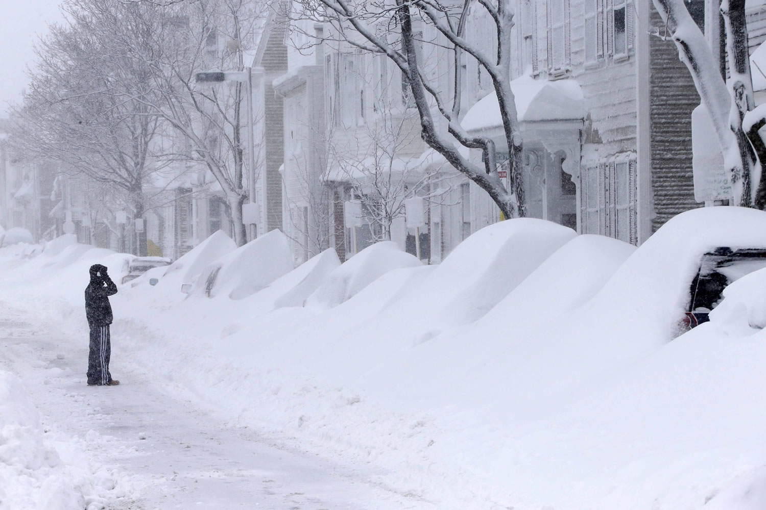 A man with a phone looks at a row of cars buried in snow Saturday in Boston.