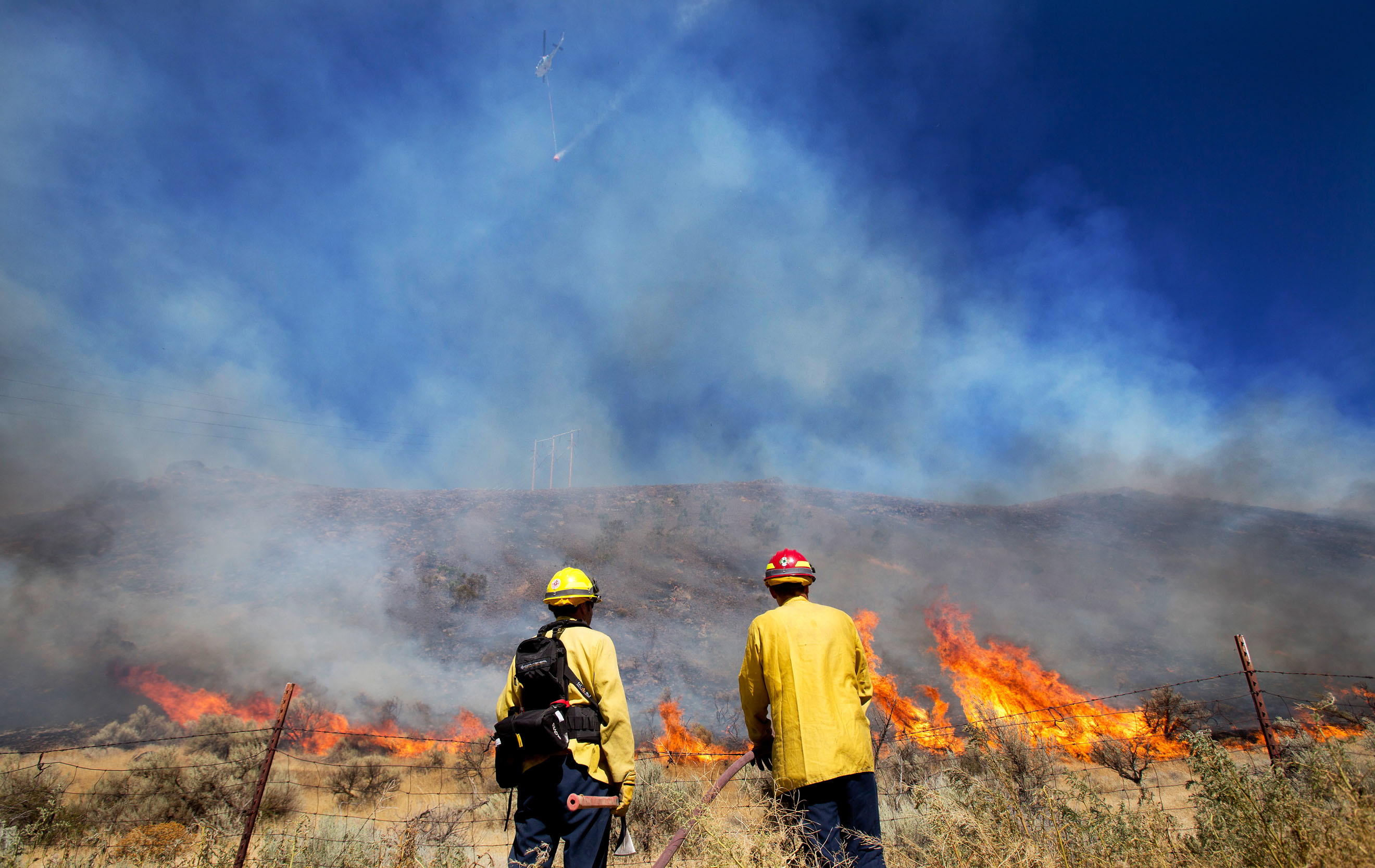 With a helicopter and water bucket flying overhead, firefighters wait for flames to reach them before spraying them down near the intersection of Clockum Pass Road and Tarpiscan Road Saturday near Malaga. Nearly 1,200 fighters are battling a wildfire that has grown to about 14 square miles near Goldendale.