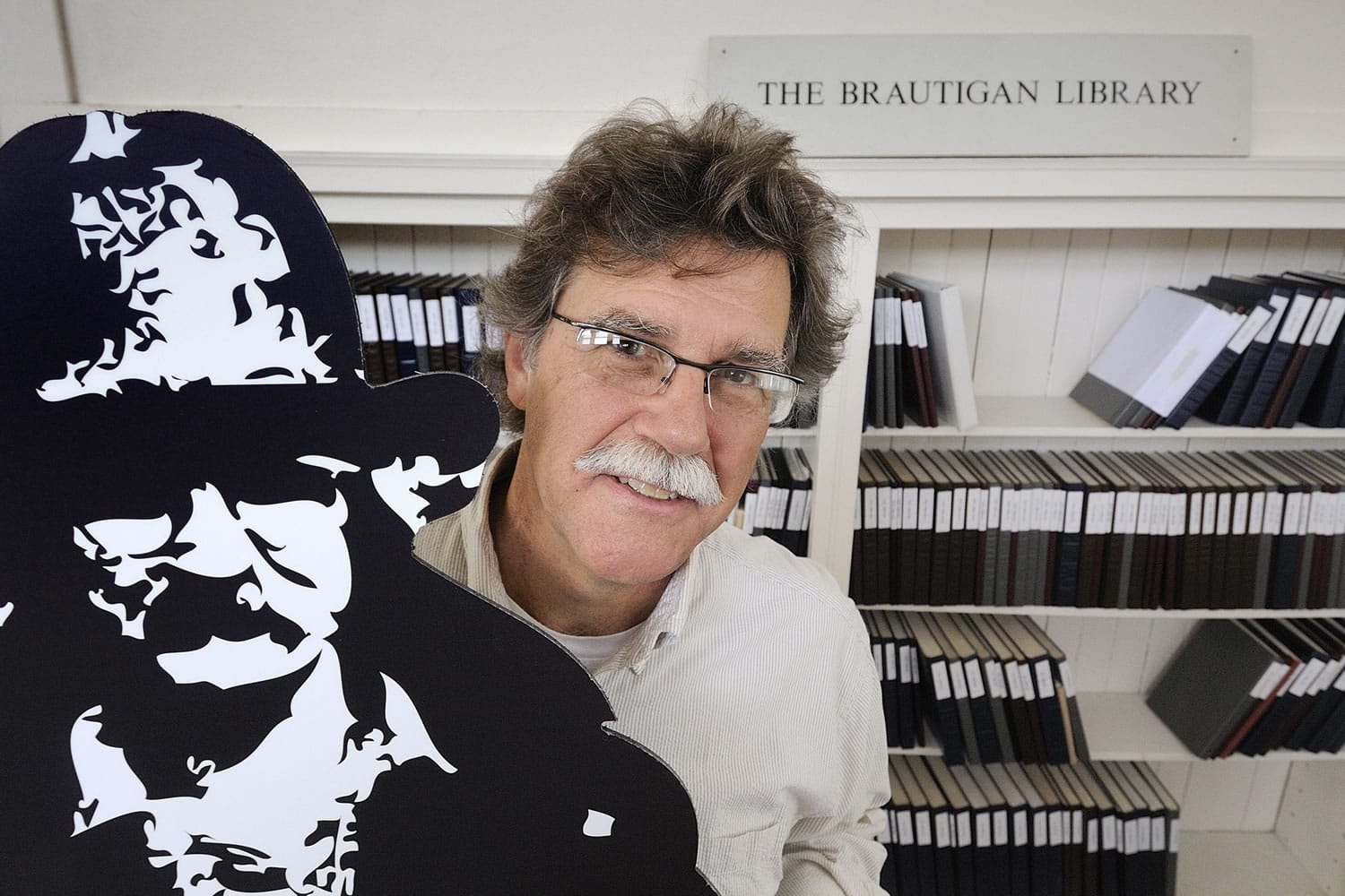 John F. Barber, with the Creative Media &amp; Digital Culture Program at Washington State University Vancouver, holds a cardboard cutout next to a display of The Brautigan Library at The Clark County Historical Museum.