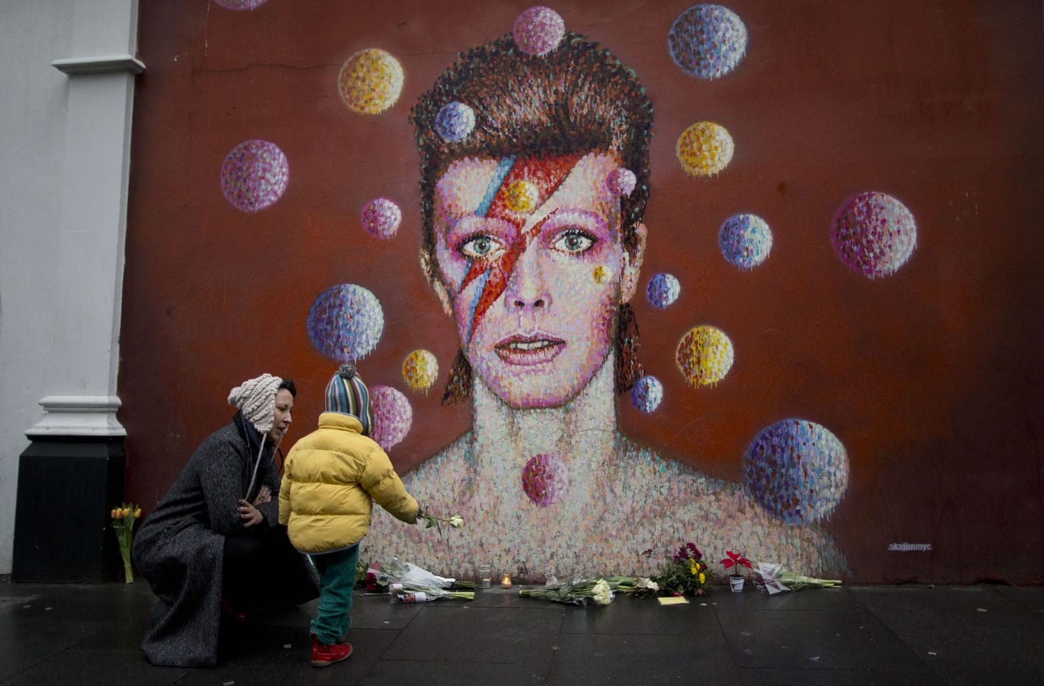 A woman and child place a flower alongside other tributes beside a mural of British singer David Bowie by artist Jimmy C in Brixton, south London, Monday, Jan. 11, 2016. David Bowie, the iconic and shape-shifting British singer whose illustrious career lasted five decade with hits like "Fame," ''Heroes" and "Let's Dance," died Sunday after a battle with cancer. He was 69.