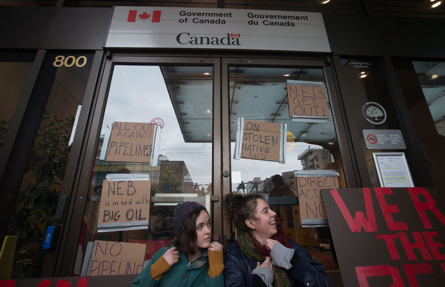 Two women sit in January outside the offices of the National Energy Board after locking themselves to the doors with bike locks around their necks, to protest the Kinder Morgan Trans Mountain Pipeline expansion, in Vancouver, B.C.