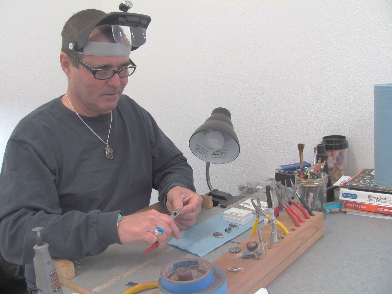 Mark Lewis uses round nose pliers to shape sterling silver wire for a sculptured pendant. He also makes earrings and bracelets, while working with semi-precious stones, Dichoric glass, fossils and cabochons.