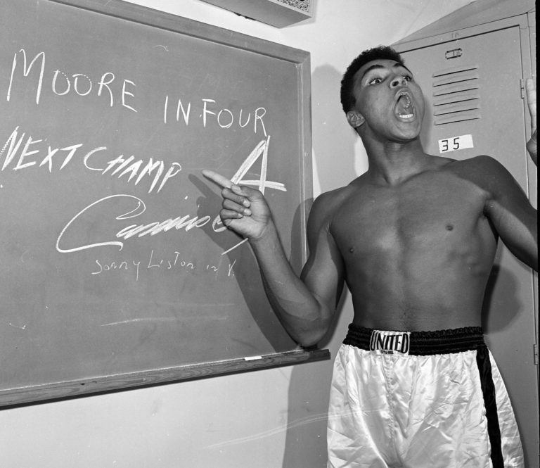 Young heavyweight boxer Cassius Clay points to a sign he wrote on a chalk board in his dressing room before his fight against Archie Moore in Los Angeles, in this Nov. 15, 1962, photo predicting he'd knock Moore out in the fourth round, which he went on to do.  The sign also predicts Clay will be the next champ via a knockout over Sonny Liston in eight rounds. (AP Photo/Harold P.