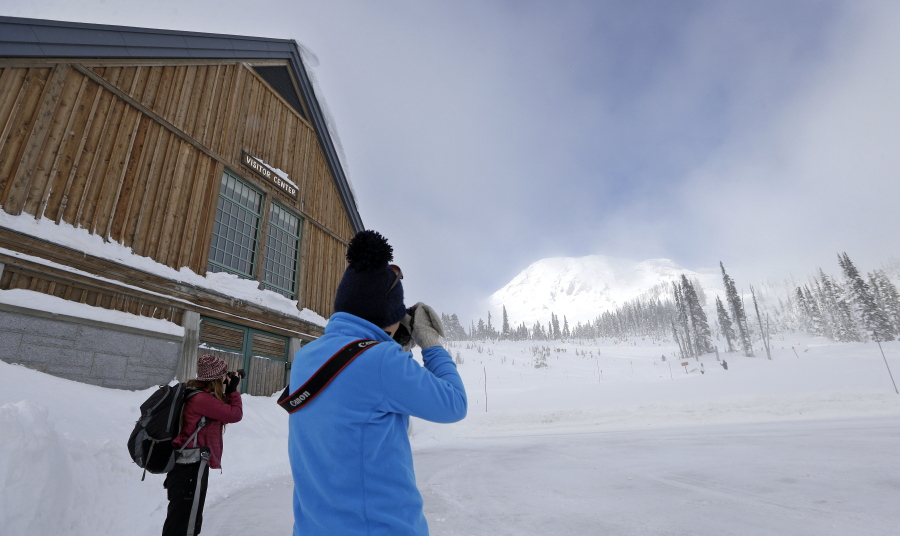 People stand outside the visitor center as they photograph Mount Rainier as it makes a brief appearance between clouds rolling in at the Paradise area at Mount Rainier National Park. Spotty or no cellular service has been the norm at the nation&#039;s fifth oldest park south of Seattle, but that could change soon. Mount Rainier National Park is considering proposals to provide cellular service in the park, setting off debates about whether people enjoying the park and surrounding wilderness areas should have access to calls and social media while in nature.