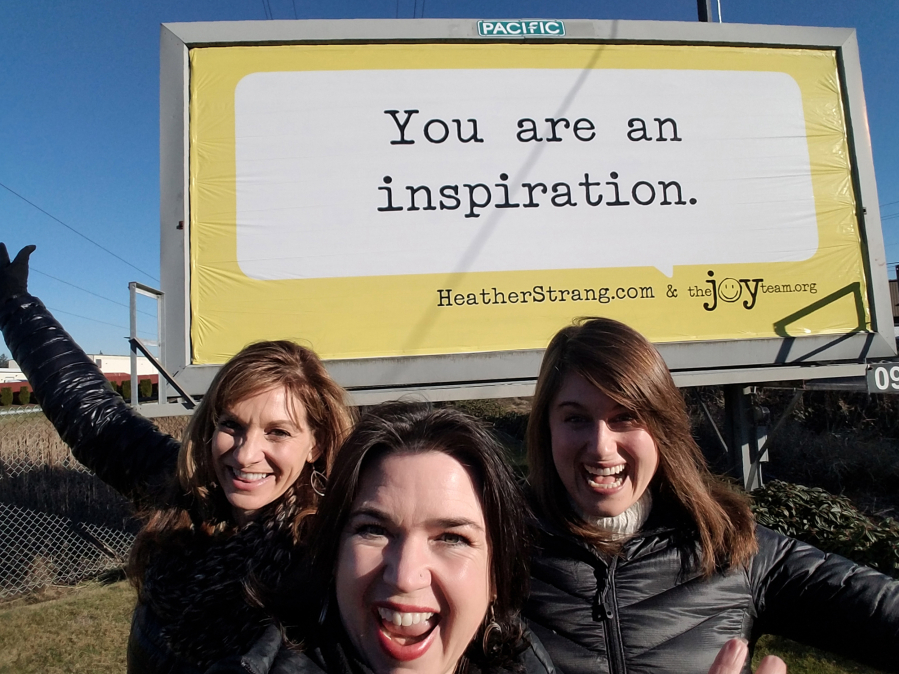 The Joy Team&#039;s Chief Joy Officer Michele Larsen, center, with co-workers Leanne Reid, left, and Megan Streeter in front of one of the organization&#039;s older billboards in Vancouver.