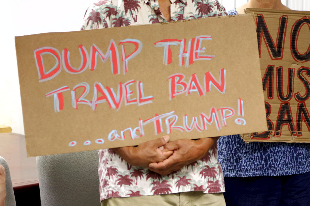 FILE- In this June 30, 2017, file photo, critics of President Donald Trump's travel ban hold signs during a news conference in Honolulu. On Tuesday, Oct. 17, 2017 a federal judge in Hawaii blocked the Trump administration from enforcing its latest travel ban, just hours before it was set to take effect. U.S. District Judge Derrick Watson granted Hawaii's request to temporarily block the policy from taking effect Wednesday.