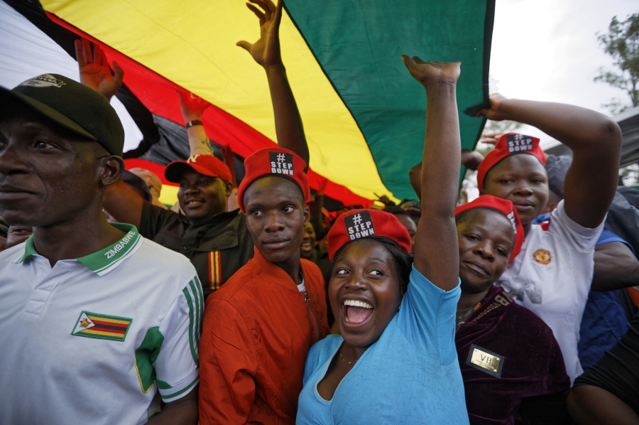 Protesters raise their fists under a large national flag, at a demonstration of tens of thousands at Zimbabwe Grounds in Harare, Zimbabwe Saturday, Nov. 18, 2017. Opponents of Mugabe are demonstrating for the ouster of the 93-year-old leader who is virtually powerless and deserted by most of his allies.