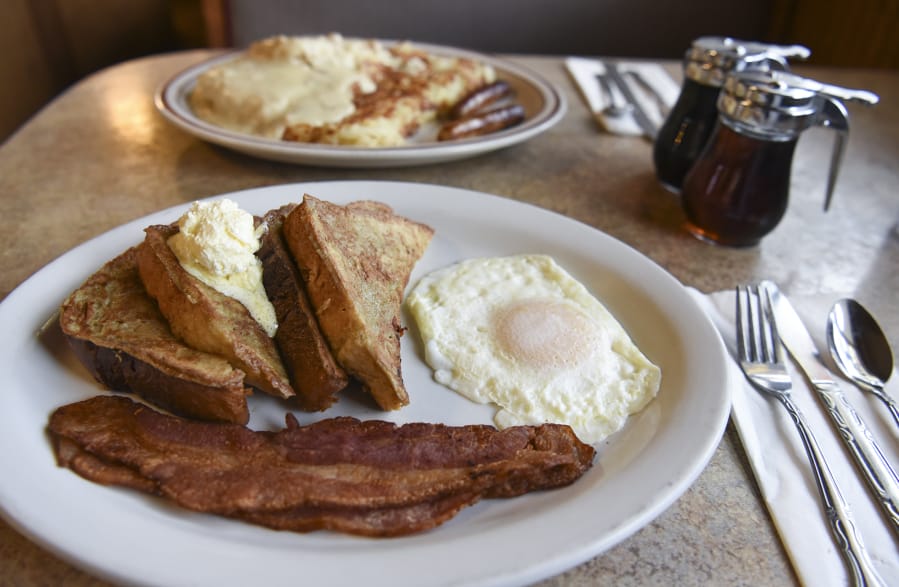 French toast with bacon and an over-easy egg are offered at The Plainsman Restaurant. Biscuits and gravy with hash browns and sausage are seen in the background.
