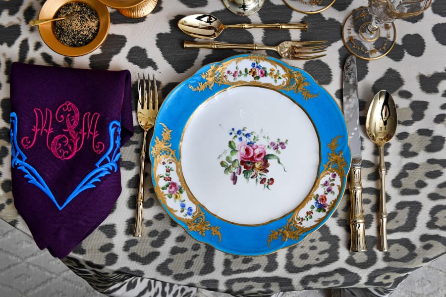 The table setting by Alex Papachristidis features Marjorie Merriweather Post’s French Sevres 1768 porcelain along with a custom tablecloth and monogrammed napkins that tie into the blues, purples and pinks of the plates. The centerpiece is a handcrafted porcelain flower arrangement by Vladimir Kanevsky. The gold salt and pepper cellars are a modern design by Christopher Spitzmiller. The bamboo-handled flatware belongs to Papachristidis.