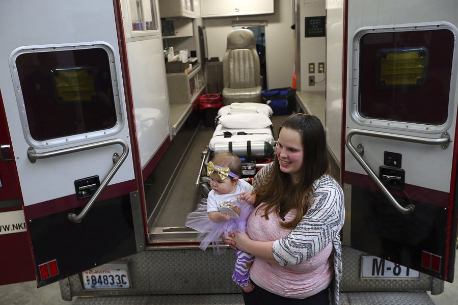 In this Tuesday, Feb. 6, 2018 photo, Lakeisha Rogers holds her one-year-old daughter Eleanor behind Medic 81 at North Kitsap Fire & Rescue Headquarters Station 81 in Kingston, Wash. Little Eleanor Rogers came into the world on Highway 307 amidst one of worst snowstorms in recent memory. She came into the world … in the back of an ambulance surrounded by grinning, weepy-eyed North Kitsap Fire and Rescue paramedics. (Meegan M.