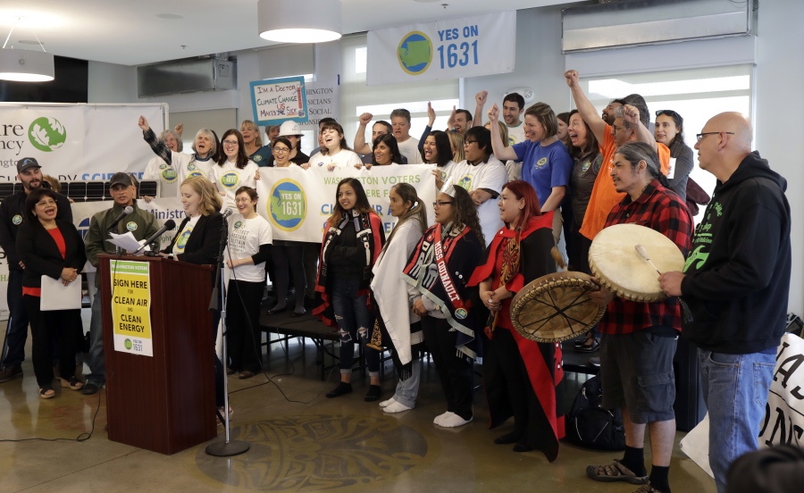 Abigail Doerr, campaign director for Yes on 1631, speaks at a podium at left, as supporters, including members of the Quinault Indian Nation at right, cheer Thursday, May 10, 2018, at a rally to kick off the campaign for “Yes on 1631,” a coalition in favor of Initiative 1631, which would levee a fee on carbon and other greenhouse-gas emissions in Washington state. If enough signatures can be gathered, the issue will go before voters in November, 2018. (AP Photo/Ted S.