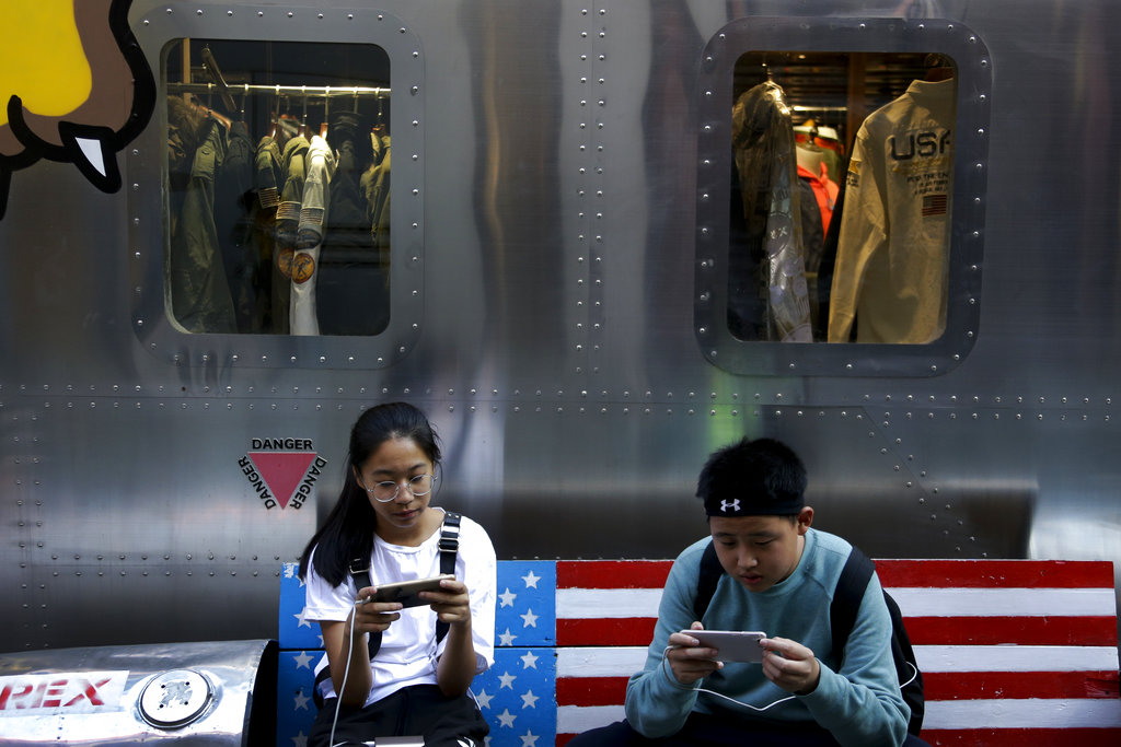 Shoppers sit on a bench with a decorated with U.S. flag browsing their smartphones outside a fashion boutique selling U.S. brand clothing at the capital city's popular shopping mall in Beijing, Monday, Sept. 24, 2018. China raised tariffs Monday on thousands of U.S. goods in an escalation of its fight with President Donald Trump over technology policy and accused Washington of bullying Beijing and damaging the global economy.