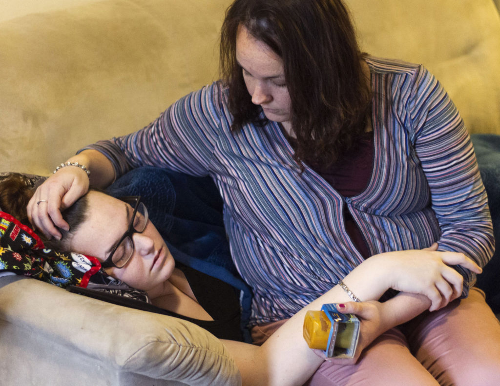 Miranda Neumann, right, embraces her 15-year-old daughter Elianna Neumann, left, recovering from her sixth round of viral meningitis, in January 2019. Miranda Neumann is holding baby food for her 6-month old.