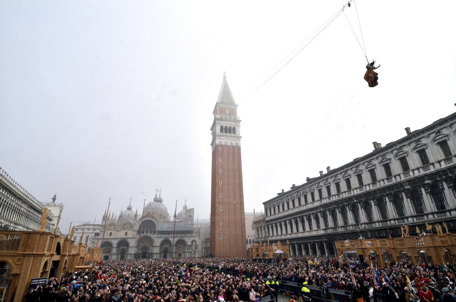 FILE - In this Sunday, Jan. 31, 2016 filer, people fill St. Mark’s Square in Venice, Italy, to watch the ‘ Flight of the Angel ‘, as a woman dressed in traditional costume descends from the bell tower into the square. The mayor of Venice says Italy’s new budget law includes a measure allowing the lagoon city to charge all visitors for accessing the historic center -- not just those sleeping in hotels or other accommodations. Mayor Luigi Brugnaro announced on Twitter late Sunday, Dec. 30, 2018 that the tax will ‘‘allow us to manage the city better and to keep it clean,’’ and ‘‘above all allow Venetians to live with more decorum.’’ The city council will determine the amount of the tax and collection mode.