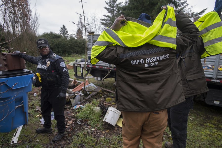 Cpl. Rey Reynolds joins employees of Rapid Response Bio Clean, a company the city contracts with to clean up homeless camps. The Vancouver Police Department previously posted a notice at the camp, telling the occupants they were in violation of the city’s camping ordinance.