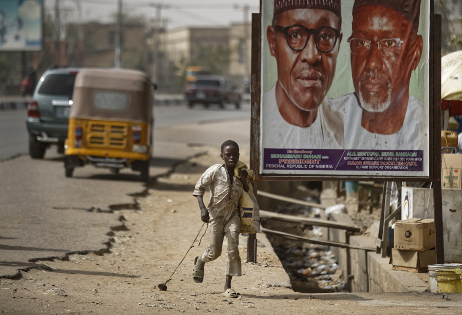 A young boy scavenges for re-sellable items from garbage on the streets, runs past a sign showing incumbent President Muhammadu Buhari, left, and local party official Mustapha Dankadai, right, in Kano, northern Nigeria Friday, Feb. 15, 2019. Nigeria is due to hold general elections on Saturday, pitting incumbent President Muhammadu Buhari against leading opposition presidential candidate Atiku Abubakar.