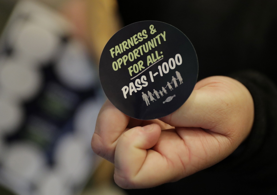 A volunteer holds a sticker showing support for Initiative 1000 outside a joint Washington state House and Senate committee, Thursday, April 18, 2019, at the Capitol in Olympia, Wash. Initiative 1000 would allow the state government to use affirmative action policies that do not constitute preferential treatment to remedy discrimination in public employment, education and contracting. (AP Photo/Ted S.