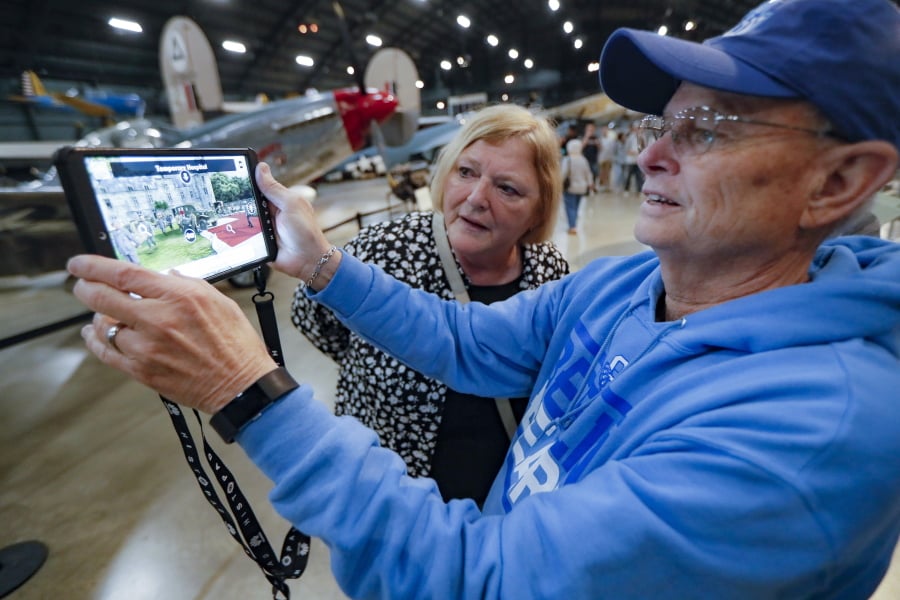 In this Friday, April 26, 2019 photo, Deane Sager, of Louisville, right, and his wife Cathy use a Histopad tablet to view scenes from operations on the western front of World War II at the The National Museum of the U.S. Air Force, in Dayton, Ohio. French-developed technology making its U.S. debut this month will allow new views of the D-Day invasion 75 years ago that began the liberation of France and helped end World War II. The National Museum of the U.S. Air Force near Dayton begins its D-Day commemorations May 13 with military-veteran paratroopers dropping from a vintage plane flying overhead, new exhibits and movies about the June 6, 1944, attack on heavily fortified German positions guarding the coastline.