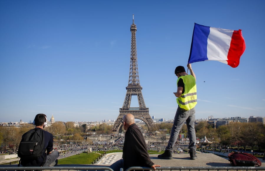 FILE - In this Saturday, March 30, 2019 file photo, a man holds a French flag as French with the Eiffel Tower in the background, during protests in Paris. Paris is wishing the Eiffel Tower a happy birthday with an elaborate laser show retracing the monument’s 130-year history.