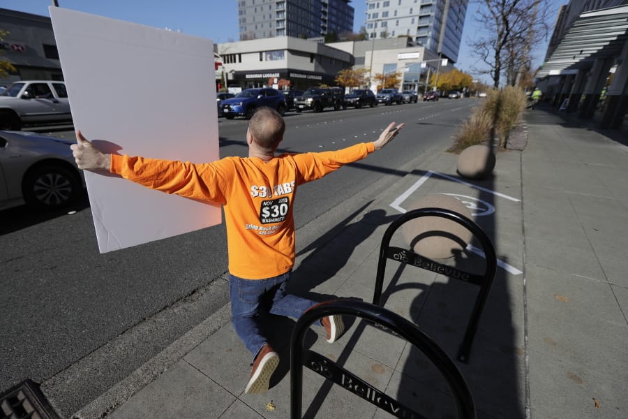 Anti-tax activist Tim Eyman kneels and begs motorists to honk their horns as he holds a sign supporting Initiative 976, which would cut most car tabs to $30 in Washington state, Tuesday, Nov. 5, 2019, on election day in Bellevue, Wash. If passed by voters, the measure would leave state and local governments scrambling to pay for road paving and other transportation projects. (AP Photo/Ted S.