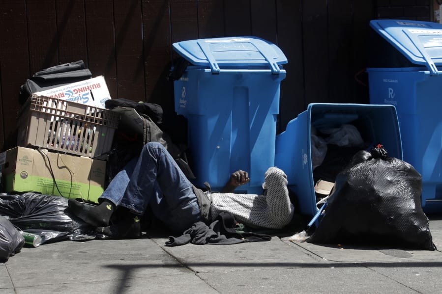 A homeless man sleeps in front of recycling bins and garbage on a street corner in San Francisco in August.