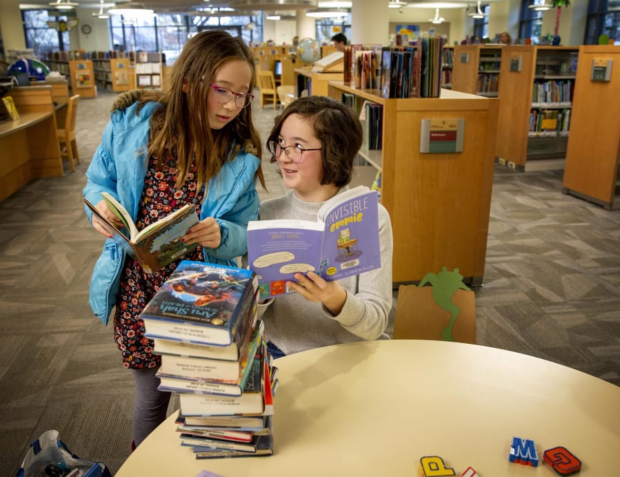 Hannah Rhee, left, listens as her sister Maya Rhee tells her about a part of the story in the book &quot;Invisible Emmie&quot; while visiting the Eugene Public Library on Thursday. Maya is a voracious reader and had a stack of books ready to check out.