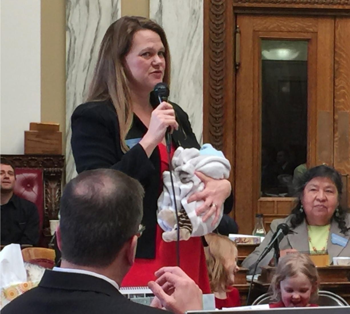 In this March 2017, photo, provided by Rep. Kimberly Dudik, Dudik speaks on the floor of the legislature holding her newborn son Marcutio in Helena, Mont. As experts predict another banner year of women running for office, hurdles remain particularly for those like Dudik who have young children. Only six states have laws specifically allowing the use of campaign funds for child care. In most states, including Montana, the law is silent on the issue and up to interpretation by state agencies or boards. (Rep. Nate McConnell/Rep.