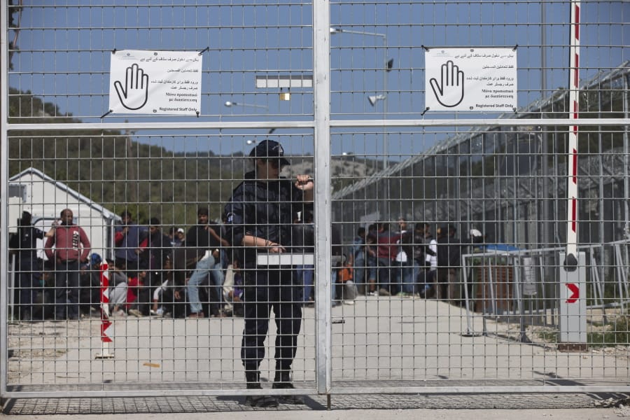 FILE - In this Tuesday, April 5, 2016 file photo, a Greek police officer closes the main gate of Moria camp as behind her refugees and migrants protest against the EU- Turkey deal about migration inside the entrance of Moria camp in the Greek island of Lesbos. Greece&#039;s government says on Monday, Feb. 10, 2020 it is planning to use emergency legal powers to create detention centers for migrants on five Greece islands to try and speed up deportations back to Turkey.