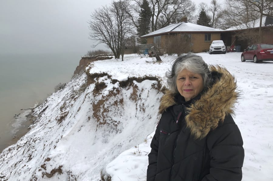 In this Jan. 14, 2020, photo, Rita Alton stands next to her house on the edge of a cliff overlooking Lake Michigan near Manistee, Mich. When her father built the 1,000-square-foot, brick bungalow in the early 1950s, more than acre of land lay between it and the drop-off overlooking the water. But erosion has accelerated dramatically as the lake approaches its highest levels in recorded history, hurling powerful waves into the mostly clay bluff.