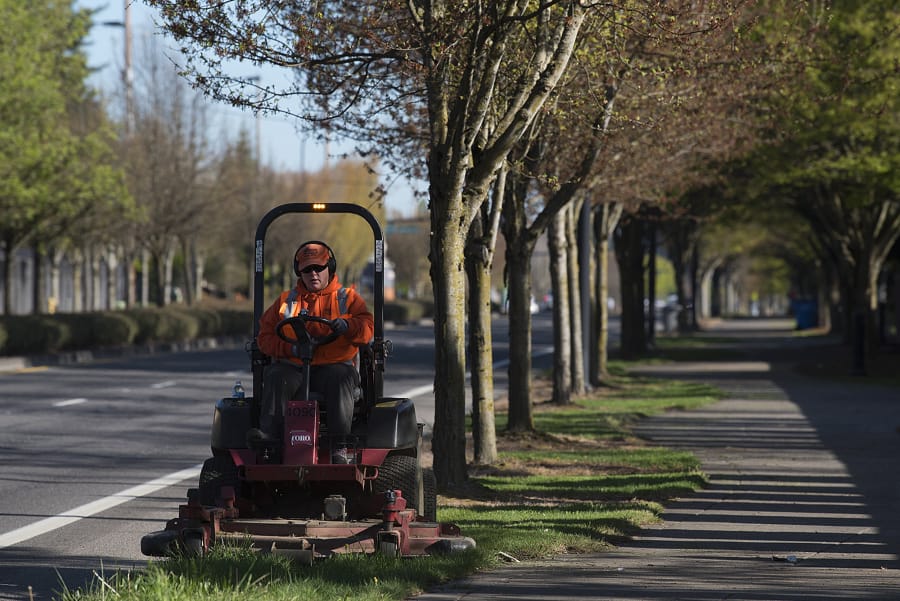 Storme Telford of the Vancouver Public Works department trims grass along Southeast 192nd Avenue on Tuesday morning. Vancouver has resumed some nonemergency public works operations, like mowing and street sweeping. Telford was given a protective mask but said she was not using it because she was away from people.