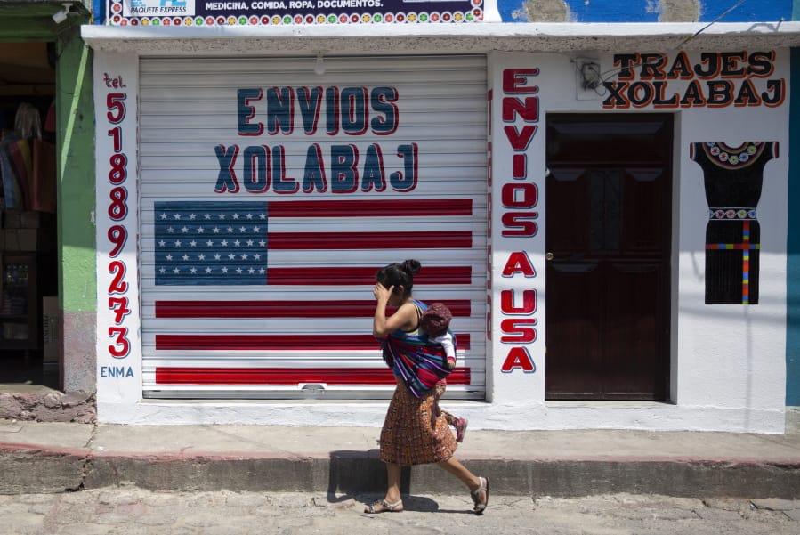 In this April 3, 2020 photo, a woman carrying a child walks past a closed courier business featuring a U.S. flag and the Spanish phrase: &quot;Send to U.S.A&quot; in the largely indigenous town of Joyabaj, Guatemala, where half of the residents depend on remittances, almost all from the U.S. The devastation wrought by COVID-19 across the developed world is cutting into the financial lifelines for people across Latin America, Africa and Asia.