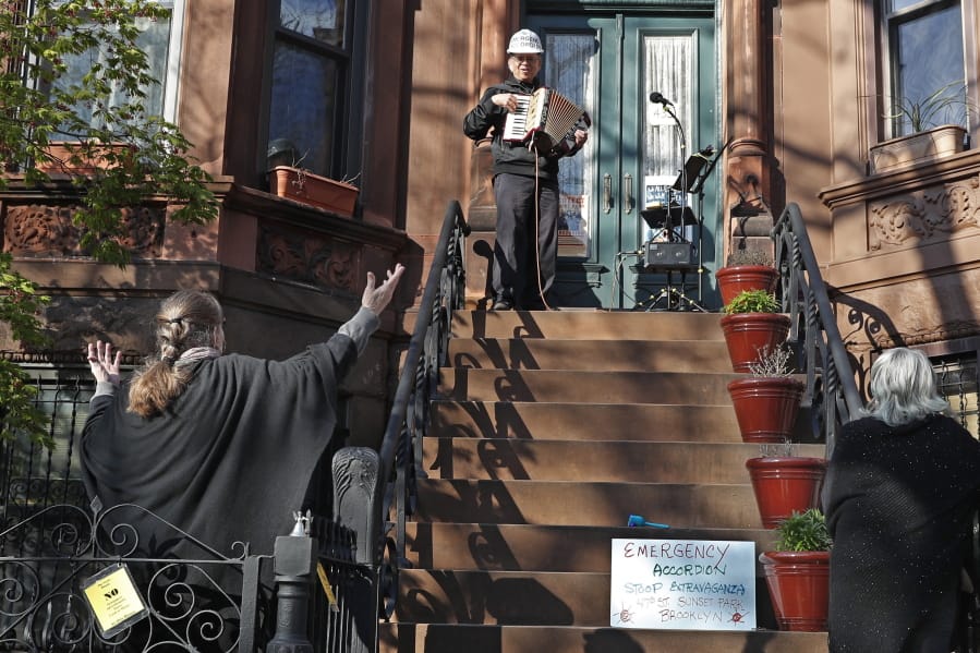 In this Wednesday, April 15, 2020 photo, choir director and neighbor Pam McAllister, left, encourages musician Paul Stein, center, to invent timely words to his songs as Stein entertains neighbors with an accordion concert from his stoop in front of his Sunset Park, Brooklyn home during the coronavirus outbreak in New York. Stein&#039;s partner Elena Schwolsky listens, lower right. &quot;I enjoy playing live music for people, and I can do that without leaving my house,&quot; Stein says. A longtime political and social activist, Stein is careful to maintain social distancing rules and encourages his neighbors to do the same. &quot;Everyone stays put in their front yards, but can still hear the music, he says. Stein uses an amplifier so neighbors down the block can also hear it.