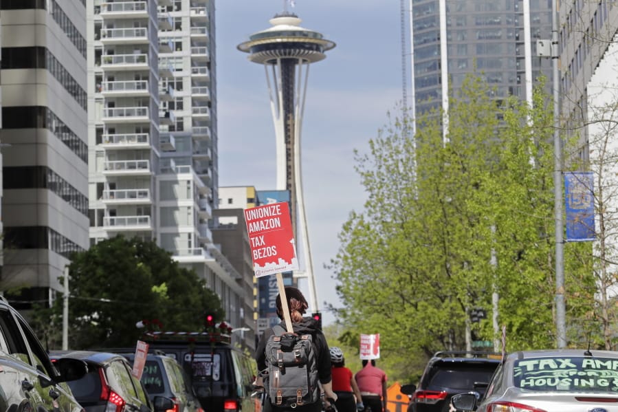 A protester carries a sign that reads &quot;Unionize Amazon Tax Bezos,&quot; while riding a bike during a car-based protest Friday, May 1, 2020, at the Amazon Spheres in downtown Seattle. May Day in Seattle traditionally brings large protests and demonstrations from many groups and causes, and this year some people stayed in their cars or otherwise tried to practice social distancing due to the outbreak of the coronavirus. (AP Photo/Ted S.