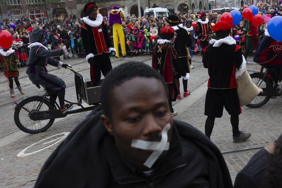 In this Sunday Nov. 17, 2013, file image, A demonstrator with a cross taped over his mouth, foreground, turns his back to the parade Black Petes, the blackface sidekicks of Sinterklaas, the Dutch version of Santa Claus, in Amsterdam, Netherlands. The death of George Floyd at the hands of Minneapolis police officers has sparked a reexamination of many countries&#039; colonial histories, actions and traditions, that often were exalted in the form of statues and other memorials. The Netherlands has been wrestling for years with issues of racism, with much of the debate revolving around the divisive children&#039;s character Black Pete, who is usually portrayed by white people wearing blackface makeup at celebrations each December marking Sinterklaas, a Dutch celebration of St. Nicholas.