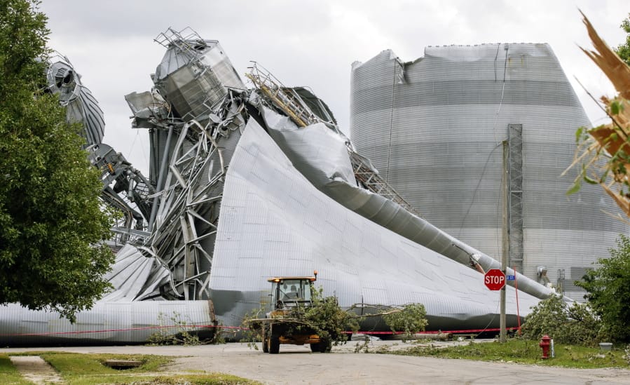 Iowa Department of Transportation workers help with tree debris removal as grain bins from the Archer Daniels Midland facility are seen severely damaged in Keystone, Iowa, on Wednesday, Aug. 12, 2020. A storm slammed the Midwest with straight line winds of up to 100 miles per hour on Monday, gaining strength as it plowed through Iowa farm fields, flattening corn and bursting grain bins still filled with tens of millions of bushels of last year&#039;s harvest.