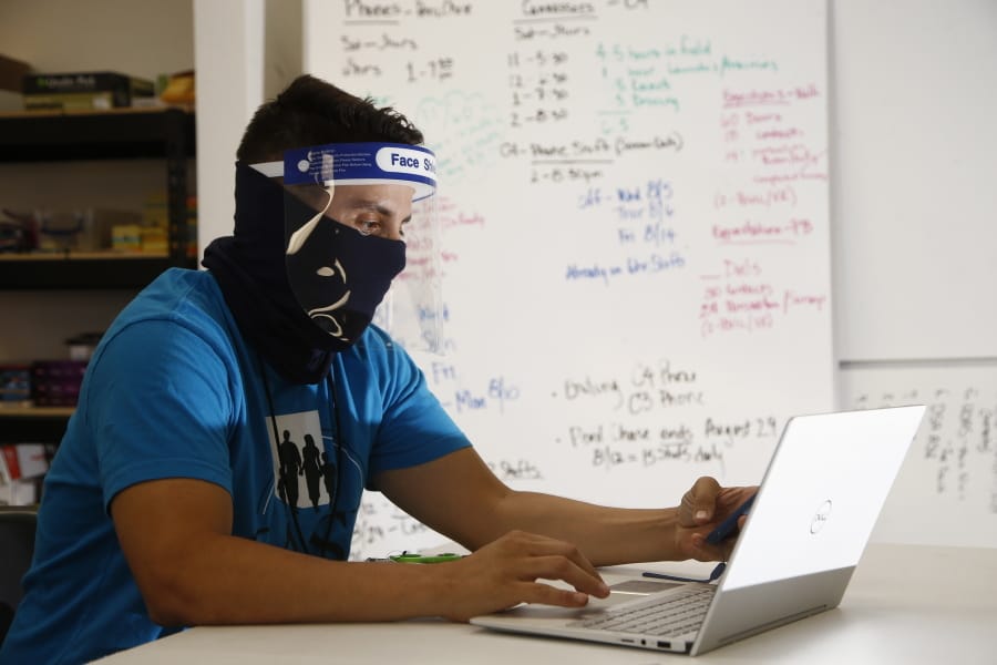 Wearing a face covering and face shield while working amid the coronavirus, Maico Olivares, lead voter registration organizer for Central Arizonans for a Sustainable Economy, works his phone and computer as he tries to reach about 25 people a day, mostly within the Latino community, to persuade them to register to vote Thursday, Aug. 6, 2020, in Phoenix. Like others who register people to vote, those efforts have become extremely difficult during the pandemic. (AP Photo/Ross D.