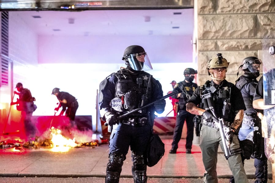 An Oregon State Police officer, right, stands watch as fellow officers extinguish a fire lit by protesters behind the Mark O. Hatfield United States Courthouse on Sun, Aug. 2, 2020, in Portland, Ore. Following an agreement between Democratic Gov. Kate Brown and the Trump administration to reduce federal officers in the city, nightly protests remained largely peaceful without major confrontations between protesters and officers.