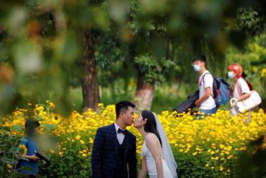 FILE - People wearing face masks pass by newlyweds kissing as they posing for wedding photos at the Olympic Forest Park in Beijing on July 2, 2020. Now that weddings have slowly cranked up under a patchwork of ever-shifting restrictions, horror stories from vendors are rolling in. Many are desperate to work after the coronavirus put an abrupt end to their incomes and feel compelled to put on their masks, grab their cameras and hope for the best.